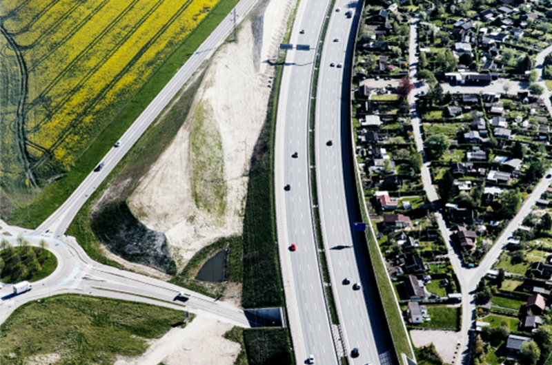 Luftfoto af en motorvej med en gul mark til højre og et boligkvarter til venstre.