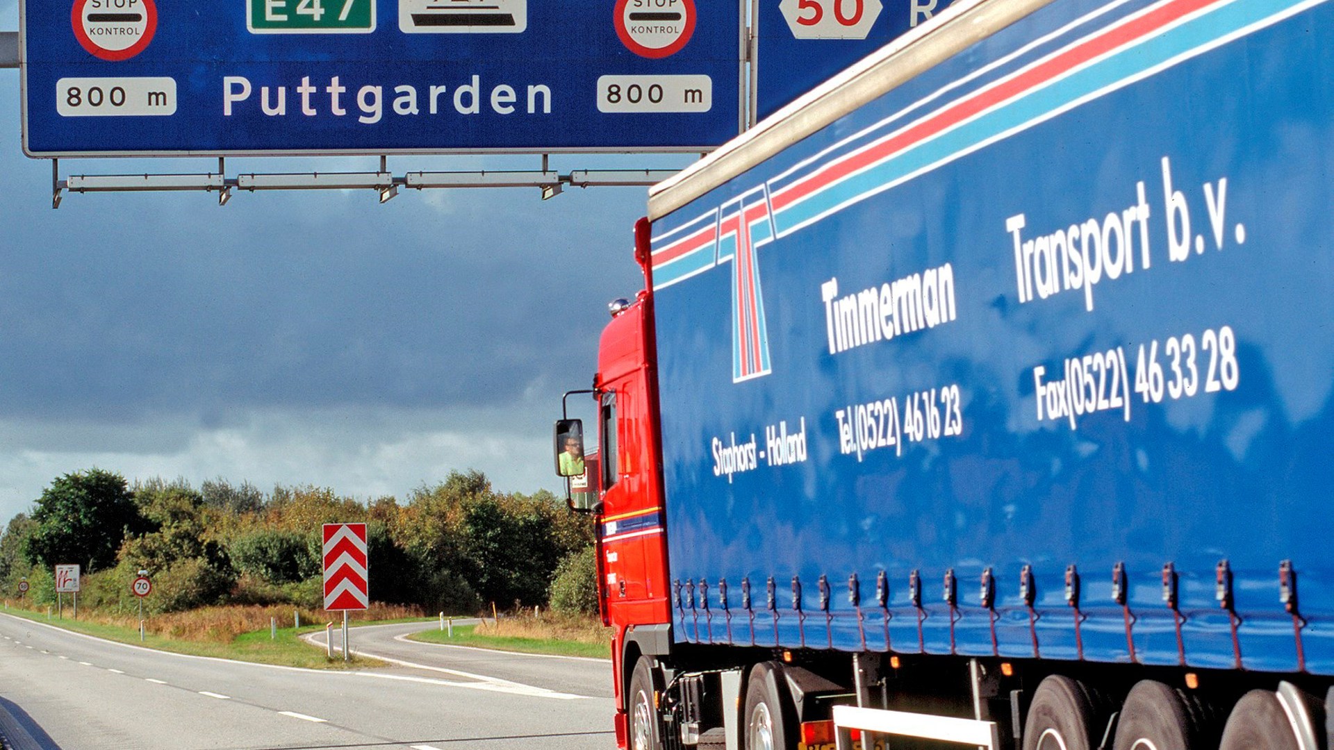 En blå lastbil kører under et motorvejsskilt hvorpå der står Puttgarden.