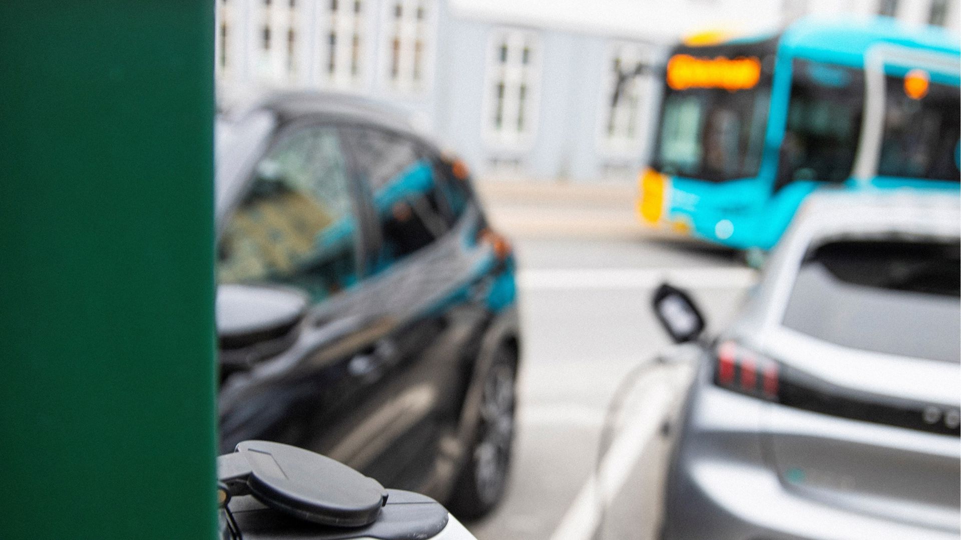 I forgrunden ses et stik til en el-bil i en grøn ladesøjle. I baggrunde en bus og to prakerede biler.