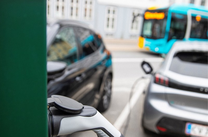 I forgrunden ses et stik til en el-bil i en grøn ladesøjle. I baggrunde en bus og to prakerede biler.