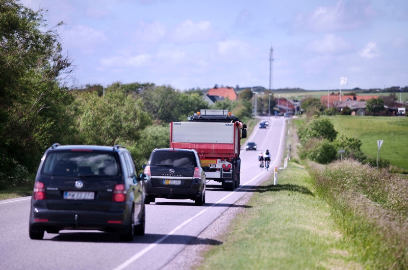 Biler og lastbil kører på landevej med cykelmarkering i siden. Længere fremme ses to cyklister og en mindre by.