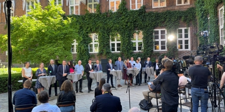 Foran en mur med grønne planter står en række mennsker ved borde. I forgrunden ses et kamera der filmer disse og ni personer der sidder med ryggen til os.